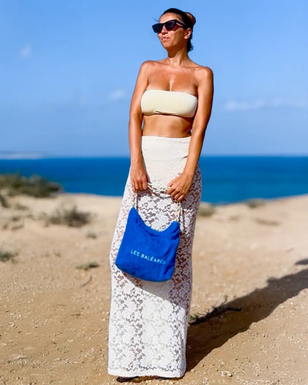 Leslie-Sulman-Beach-Beyond-Collection-26-baleares-royal-blue Mannequin brune face à la mer Méditerranée. Elle porte une jupe longue en dentelle transparente blanche, un haut de maillot de bain forme bandeau crème.Elle porte un sac de plage personnalisé de la marque Leslie Sulman à la main. Il est de couleur royal blue et il est brodé du message Les Baléares en blanc.